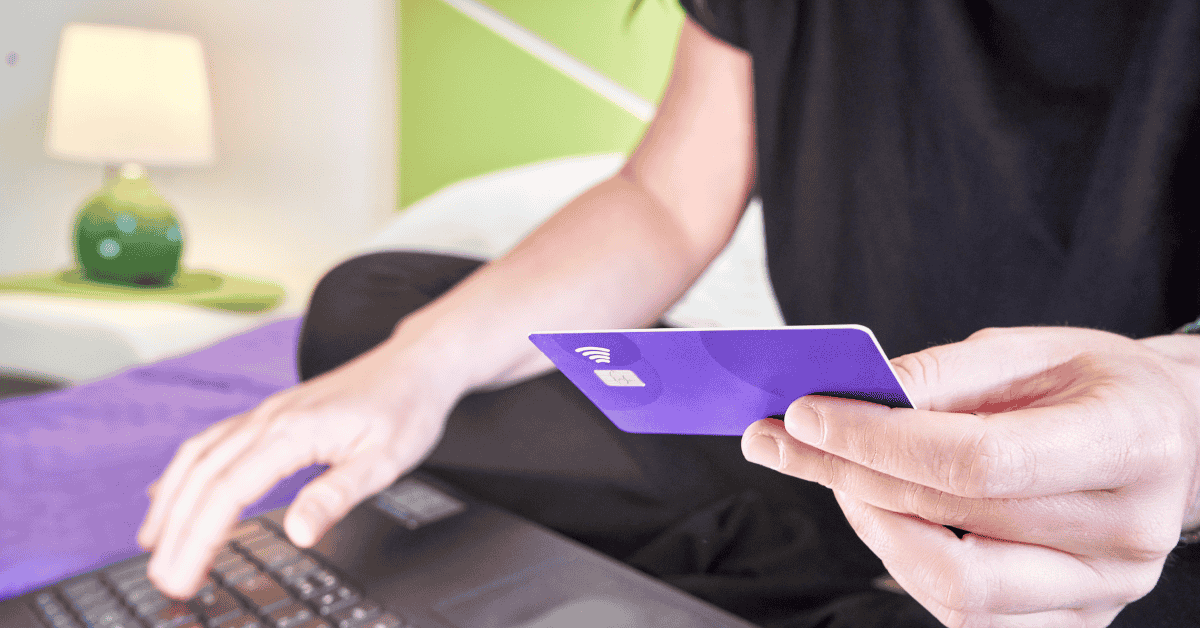 Persona usando una tarjeta de crédito morada para hacer una compra online desde su laptop en casa.