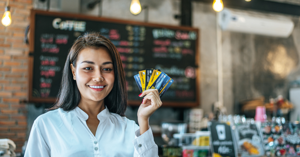 Mujer sonriente mostrando varias tarjetas de crédito en un café, representando opciones de financiamiento y consumo inteligente.