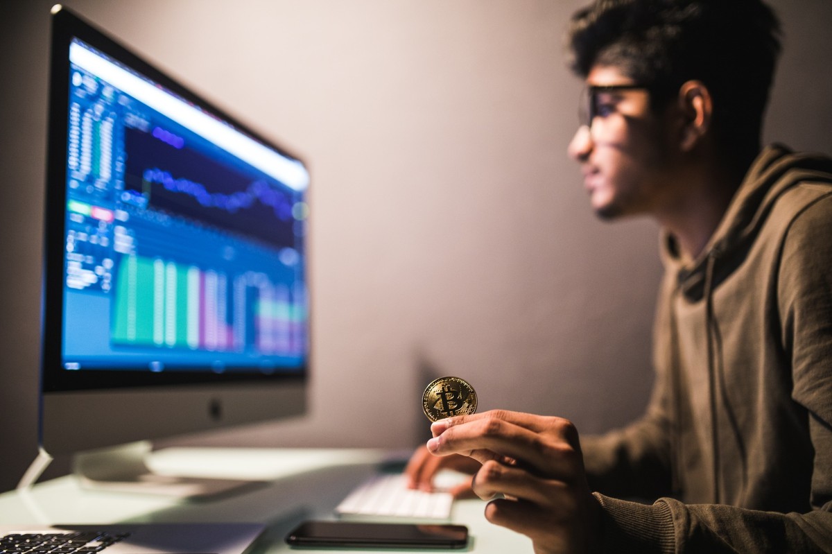 Un joven sentado frente a una computadora en un entorno de oficina analizando gráficos financieros en pantalla mientras sostiene una moneda física de bitcoin. La escena transmite concentración y el concepto de inversión digital y criptomonedas