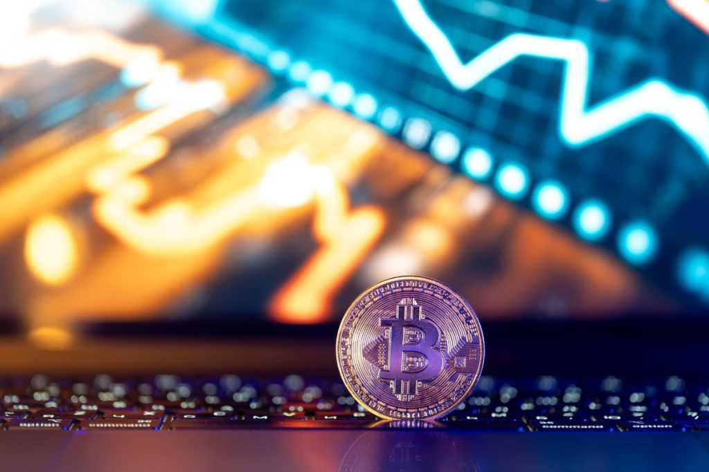 Una moneda de bitcoin dorada colocada sobre un teclado de computadora, con un fondo desenfocado que muestra gráficos de mercado y luces azules y naranjas, simbolizando el dinamismo y la volatilidad del mercado de criptomonedas.