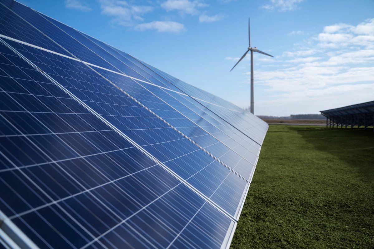 Fotografía de paneles solares brillando bajo el sol en un campo verde, con un aerogenerador visible al fondo, simbolizando la integración de energía solar y eólica en la transición hacia fuentes renovables sostenibles.