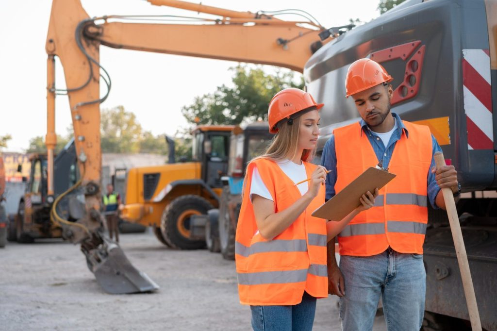 Dos ingenieros o trabajadores de la construcción con cascos y chalecos naranjas revisando planos en una obra, con maquinaria pesada al fondo. Representa la infraestructura y obra pública como motor de inversión y desarrollo económico.